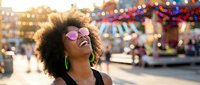 Young African American woman with curly afro hair wearing sunglasses laughing joyfully at colorful amusement park with carousel and bright lights in background.