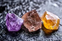 Three raw gemstone crystals in purple, brown and yellow colors arranged on dark wet stone surface for jewelry making and mineral collection purposes.