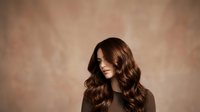 Young woman with long, wavy brunette hair and fair complexion wearing dark clothing, photographed against neutral beige studio background for beauty and fashion use.