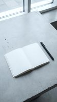 Open notebook with blank pages and black pen on concrete desk near bright window, suitable for business planning, creative writing, and office workspace concepts.