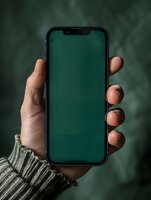 Hand holding modern smartphone with green screen display, wearing gray knit sweater against blurred emerald background. Mockup ready for product placement.