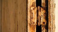 Rusty metal door hinge on weathered wooden frame showing age and corrosion. Vintage hardware detail with peeling paint and natural wood grain texture.
