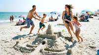 Family playing around sandcastle on sunny beach with ocean and other beachgoers in background, summer vacation fun and outdoor recreation.