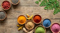 Colorful superfood powders in wooden bowls on rustic table with turmeric root, goji berries, chia seeds, and fresh green leaves.