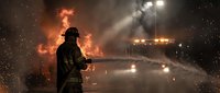 Firefighter in protective gear battles intense blaze with water hose during nighttime emergency response operation with dramatic flames and smoke.