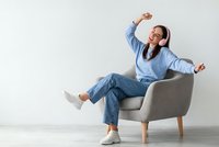 Young Asian woman listening to music with headphones dancing in modern armchair wearing casual blue outfit against white wall background for lifestyle content.