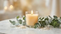 Burning candle in glass holder surrounded by eucalyptus branches and sea salt on marble surface creating peaceful spa atmosphere for relaxation and wellness.