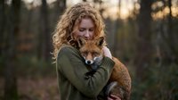 Young Caucasian woman with curly blonde hair embracing red fox in forest during golden hour sunset creating warm intimate wildlife connection moment.