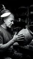 Senior male potter crafting ceramic vessel in traditional workshop under focused lamp light. Monochrome artisan at work creating handmade pottery with skilled hands.