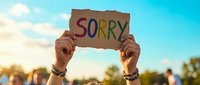 Hands holding cardboard sign with sorry message against blue sky background for apology and forgiveness concepts in outdoor setting.