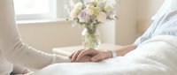 Caring visitor holds the hand of a patient resting in a hospital bed, with soft flowers in the background, conveying comfort and support.