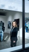 Professional woman with long brown hair, Caucasian ethnicity, wearing black business blazer and pants, standing inside modern art gallery with contemporary artwork displayed on white walls, viewed through large glass window with black frame.