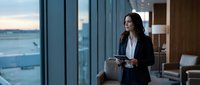 Professional businesswoman with tablet standing by office windows overlooking city skyline at dusk, corporate executive planning strategy in modern workplace.