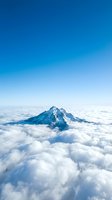 Snow-capped mountain peak rising above dense cloud layer under clear blue sky, aerial view for travel and nature backgrounds.