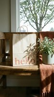 Kraft paper shopping bag with hello text on rustic wooden bench near window, green plant and terracotta blanket in warm natural light.