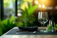 Elegant restaurant table setting with wine glasses against blurred green plant background, creating warm ambiance for fine dining experience.