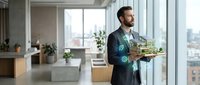 Bearded male professional in dark business suit holding architectural model with digital holographic overlays in modern office with city skyline view.
