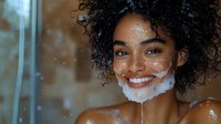 Young woman with dark curly hair and medium skin tone applying exfoliating facial scrub, smiling at camera in bathroom setting with water droplets and warm lighting.
