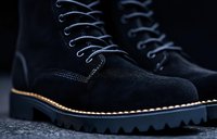 Black suede boots with contrast stitching and chunky rubber sole featuring decorative welt detail. Close-up view highlights premium materials and craftsmanship.