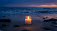 Glowing candle in glass jar on sandy beach at twilight with ocean waves and colorful sunset sky creating peaceful meditation atmosphere for relaxation.