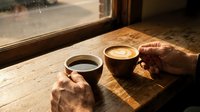 Two hands holding coffee cups on wooden table by window with warm morning sunlight creating cozy cafe atmosphere for relaxation and conversation.