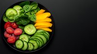 Fresh colorful vegetables and berries arranged on black plate including cucumber slices, yellow peppers, raspberries, avocado and baby spinach leaves on dark background.