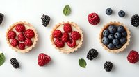 Fresh berry tartlets with cream filling arranged in row, raspberries, blueberries and blackberries scattered around with mint leaves on white background.