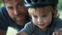 Young child wearing bicycle helmet smiles at camera while adult male figure watches protectively in background, capturing family bonding during outdoor activities.