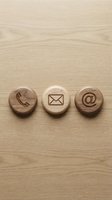 Three wooden circular buttons with contact icons - phone, envelope, and email symbol - arranged on light wood surface for business communication concepts.