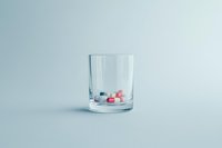 Assorted pills and capsules inside a clear glass on a light blue background, symbolizing medication, addiction, or healthcare concepts.