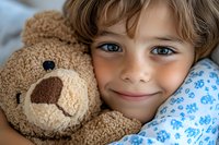 Adorable young boy with curly blonde hair cuddling brown teddy bear in bed, showing innocent smile and bright blue eyes. Childhood comfort and bedtime concept.