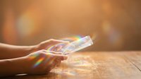 Young adult hands holding transparent crystal prism refracting rainbow light spectrum onto wooden surface, creating colorful optical effects for science, education, and creative design projects.