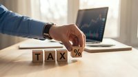 Male hand placing wooden block with percent symbol next to tax text on desk with laptop displaying financial charts in background, representing tax calculation, financial planning, and accounting services.