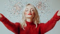 Young blonde woman in red sweater with closed eyes and red lipstick enjoying moment while glitter confetti falling around her against light background.