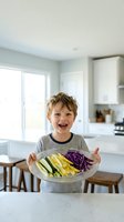Happy young boy holding plate of fresh colorful vegetables in modern kitchen smiling with excitement for healthy eating and cooking activities.