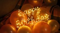 Glowing happy birthday wire light sign surrounded by golden orange balloons in warm bokeh, festive party decoration for celebration.