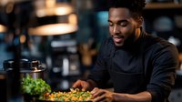 Young African American male chef in black uniform preparing fresh salad in modern restaurant kitchen. Professional culinary expert cooking healthy meal.
