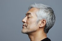 Profile view of mature Asian man with gray hair and closed eyes wearing black against gray background, expressing serenity and mindfulness.