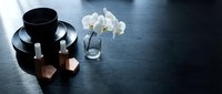 Elegant black coffee cup with saucer, white orchid flowers in glass vase and geometric copper decorations on dark wooden table surface with copy space.