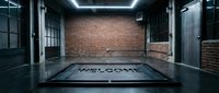 Industrial garage interior with brick wall, glowing ceiling lights, and a welcome mat on dark concrete floor, creating a moody urban atmosphere.