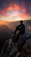 Young man sitting on mountain cliff edge watching dramatic sunset over canyon landscape with flying birds and colorful sky for adventure travel concepts.