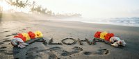 Aloha written in wet sand on a tropical beach at sunset, decorated with colorful hibiscus flowers and seashells, warm golden light.