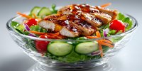 Grilled chicken breast slices with teriyaki glaze on fresh mixed greens salad with cherry tomatoes and cucumber in glass bowl for healthy dining.