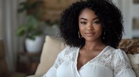 Young African American woman with natural curly hair wearing white lace dress smiling at camera while sitting on beige couch in cozy home interior setting.