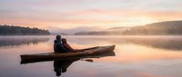 Two people sitting in wooden canoe on calm misty lake at sunrise, peaceful water reflection and forested shoreline, serene outdoor adventure and nature exploration.