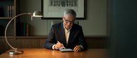 Mature male professional with gray hair and glasses wearing dark business suit sitting at wooden desk writing with pen under arc lamp in modern office environment.