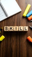 Wooden blocks spelling skill surrounded by colorful markers and open notebook on wooden desk for education, training, and professional development concepts.