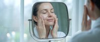 Woman with fair skin applying skincare cream to her face while looking in bathroom mirror, demonstrating daily beauty and skincare routine.