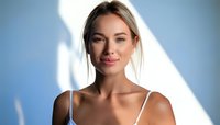 Young blonde woman with natural makeup smiling confidently against blue wall with soft shadow patterns, wearing white straps.