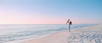 Young woman in white dress walking barefoot on pristine sandy beach at sunset with calm ocean waves and pastel sky colors creating peaceful coastal scene.